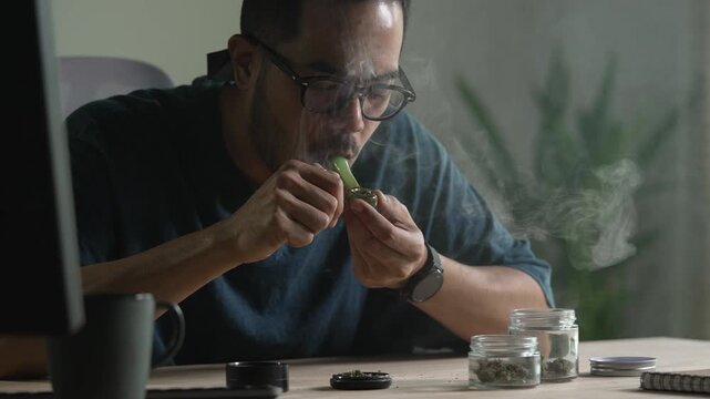 Man smoking a cannabis from smoking pipe, with cannabis equipment and accessories on the table.