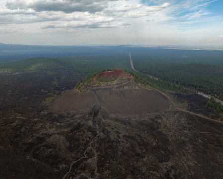 Aerial view of the Lava Butte cinder cone showing its red crater rim and dark volcanic slopes surrounded by vast lava fields and pine forests in Deschutes County, Oregon, United States.