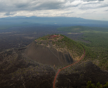 Aerial view of Lava Butte cinder cone surrounded by dark volcanic fields and evergreen forests under a dramatic cloudy sky in Deschutes County, Oregon, United States.