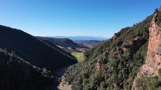 A cinematic 4K drone shot looking down a rugged mountain valley near Khenifra, Morocco; featuring steep limestone cliffs, dense cedar forests, and a distant view of lush green pastures under a clear b
