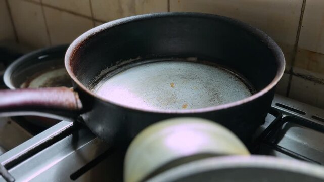 Close-up of an empty black saucepan on a gas stove in a worn indoor kitchen, with stained tiles, metal burners, and used cookware in soft natural light.