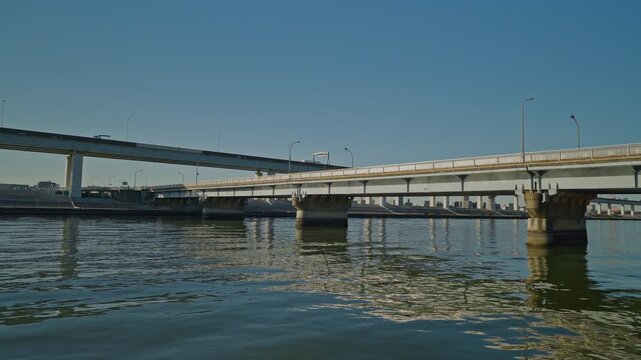 A sweeping wide shot from the water level showing several elevated expressways and bridges spanning a wide river. The camera pans across the industrial landscape under a bright, cloudless sky.