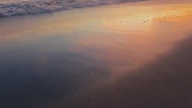 Colorful sunset sky reflects on wet sand as gentle shallow waves roll onto the shore and recede. Peaceful beach scene with soft motion and vivid coastal light.