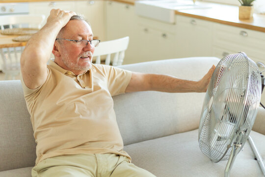 Senior man refreshing himself near fan during summer heat wave hot day. Elderly man suffering from summer heat trying to survive with electric fan. Man enjoying refreshing air blowing from ventilator
