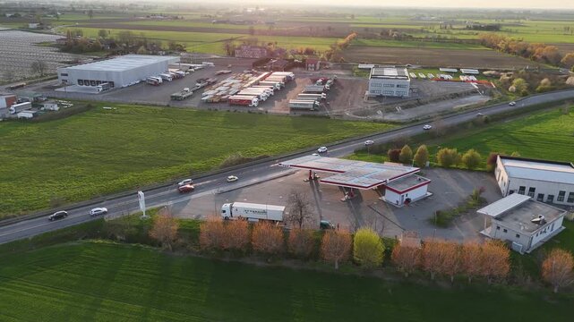 Traffic flows in Mixed landscape near Alseno Emilia Romagna Italy along SS9 Via Emilia with fuel station, parked trucks in logistics depot, low tunnel agricultural fields in Po Valley, lateral pan 180