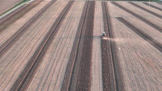 Tractor performing soil tillage in Pianura Padana near Piacenza Emilia Romagna Italy forming long parallel furrows and dust trail across agricultural field, slow drone descent shot
