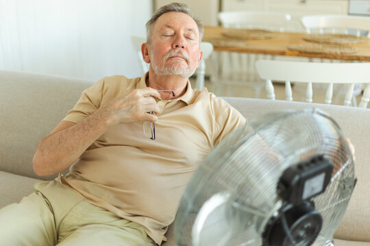 Senior man refreshing himself near fan during summer heat wave hot day. Elderly man suffering from summer heat trying to survive with electric fan. Man enjoying refreshing air blowing from ventilator