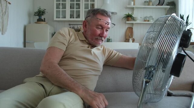 Senior man refreshing himself near fan during summer heat wave hot day. Elderly man suffering from summer heat trying to survive with electric fan. Man enjoying refreshing air blowing from ventilator