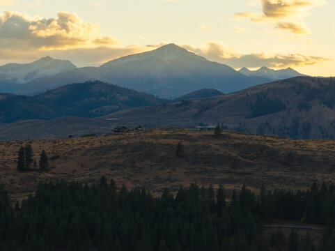 Aerial view of rolling hills, dense evergreen forests, and majestic mountain peaks under a golden sunset sky in Winthrop, Washington, United States.
