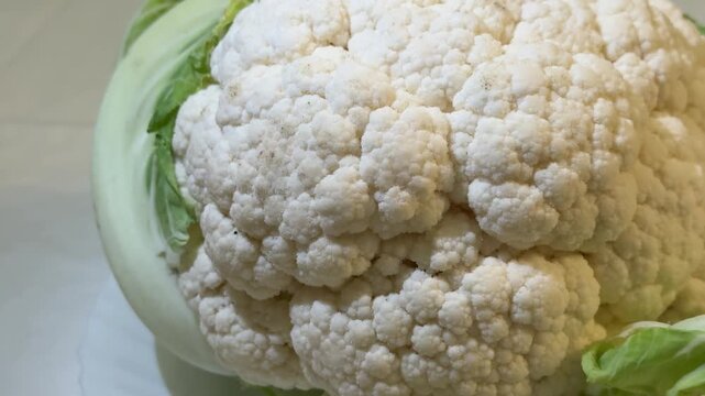 Cauliflower on white plate close-up, pristine curd and pale green leaves, soft studio lighting reveals granular texture and natural.