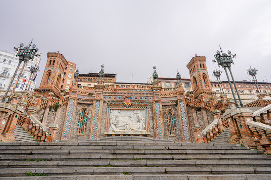 The historic Escalinata del Ovalo staircase in Teruel, Spain, featuring intricate Mudejar style brickwork and stone sculptures under a cloudy sky.