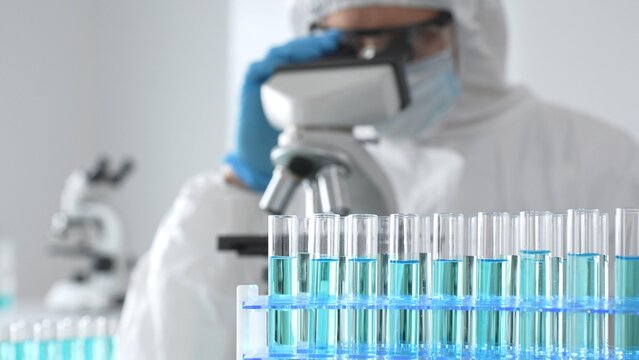 A medical virologist in protective gear and blue gloves examines viral samples under a microscope while conducting research in a sterile laboratory setting