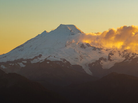 Aerial view of the snow-capped peak of Mount Baker illuminated by golden sunset light with clouds drifting across the slopes in Whatcom County, Washington, United States.