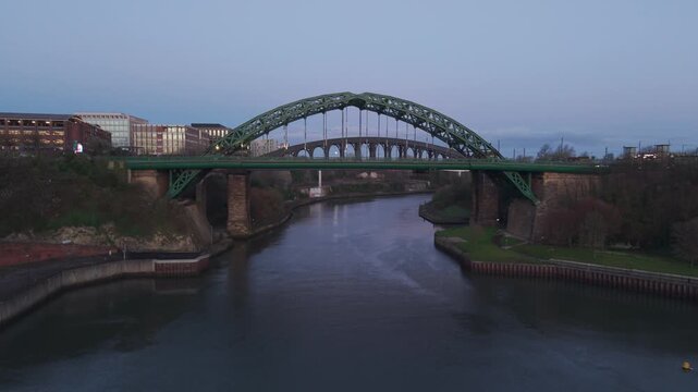 Aerial drone shot flying upwards and backwards from the Wearmouth Bridge and Monkwearmouth Railway Bridge in Sunderland at dawn, revealing the river and city buildings in twilight.