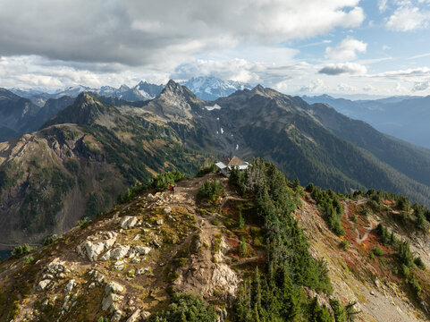 Aerial view of Winchester Mountain Lookout on a rugged peak with hikers nearby and surrounding mountain ranges under a cloudy sky in Whatcom County, Washington, United States.