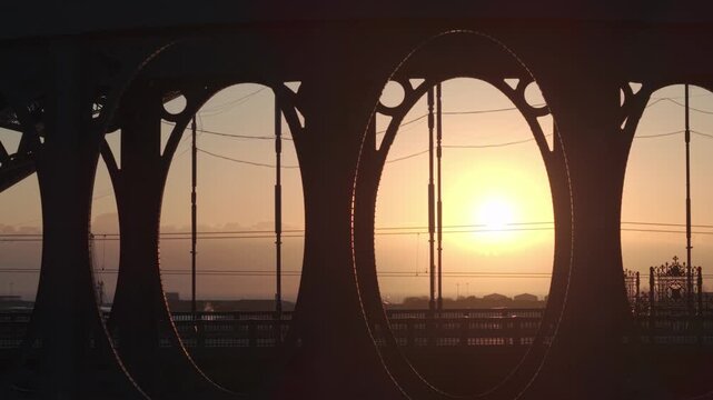 Fast aerial tracking shot of the Wearmouth and Monkwearmouth Railway Bridges in silhouette against a golden sunrise in Sunderland, featuring commuters and city traffic.
