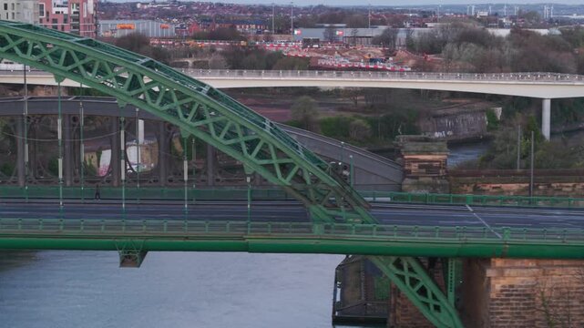 Aerial telephoto tracking shot moving laterally past the Wearmouth Bridge in Sunderland at dawn, showcasing early morning traffic commuting over the River Wear with industrial architecture in the back