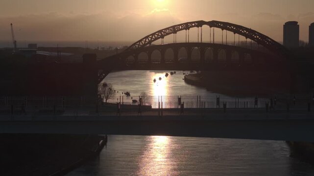 Aerial drone shot flying backwards and ascending from the silhouetted Keel Crossing, revealing the Wearmouth Bridge and Monkwearmouth Railway Bridge against a golden sunrise in Sunderland.
