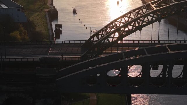 Aerial telephoto tracking shot of a lone pedestrian crossing the Wearmouth Bridge at sunrise in Sunderland, with the industrial architecture of the Monkwearmouth Railway Bridge in the foreground.