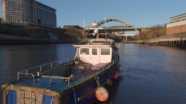 Low-altitude aerial tracking shot passing a small weathered boat moored on the River Wear, with the Wearmouth Bridge and Monkwearmouth Railway Bridge in the background in Sunderland.