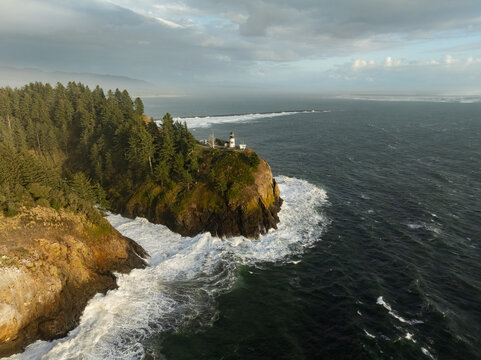 Aerial view of Cape Disappointment Lighthouse perched on a rocky cliff with crashing waves and a dense evergreen forest in Pacific County, Washington, United States.