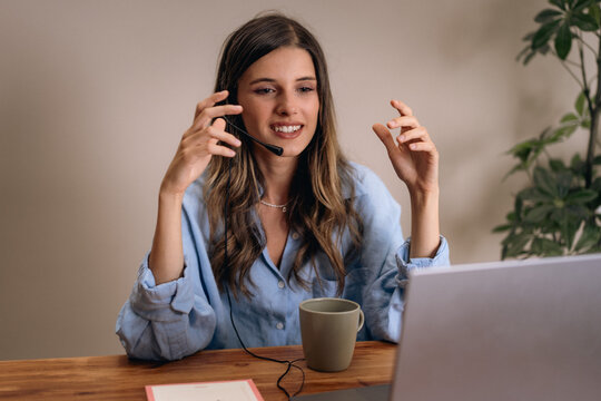Woman in online meeting with headset smiling while working remotely from home. Modern workspace with woman engaging in digital collaboration via laptop and headset.