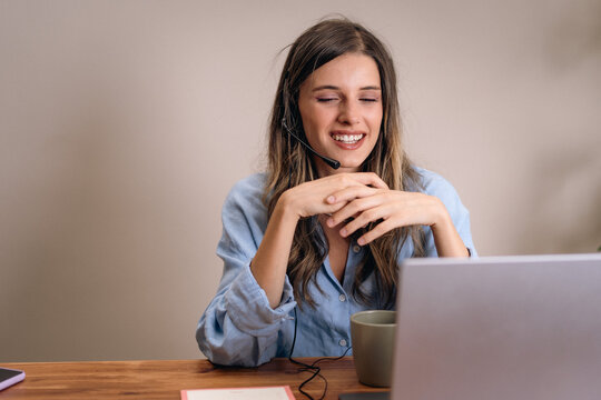 Happy woman on video call wearing headset during remote work from home. Young woman enjoying a virtual meeting with colleagues, wearing a headset and smiling at the laptop.
