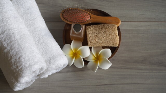The image depicts a wellness spa atmosphere, featuring rolled-up white towels, essential oils, hairbrushes, loofahs, and frangipani flowers on a wooden table, conveying the concept of relaxation thera