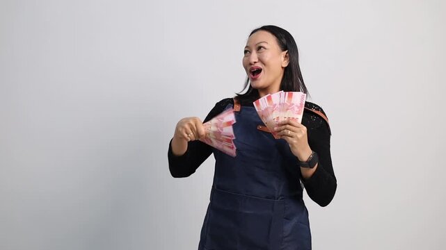 Asian woman in blue apron dancing happily while holding some rupiah banknotes, profit or salary from business.