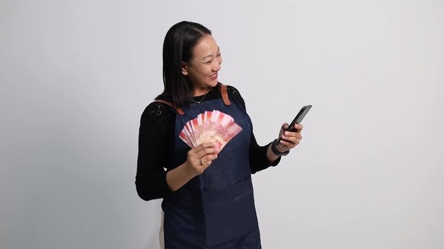 Asian woman wearing an apron with a surprised expression while using his cellphone and carrying rupiah banknotes, happy to get profits from selling online.