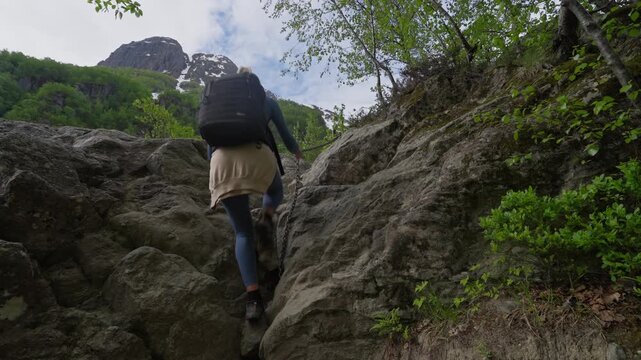 Adventurous hikers climb a steep rock wall using a safety chain, with a snow-capped mountain peak visible in the distance. Thrilling alpine ascent in rugged natural scenery