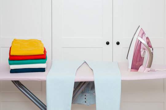 Clean colorful female jeans and electric iron with steam system on ironing board at white wardrobe at home room. Closeup. Front view.
