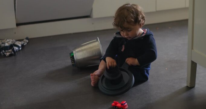 Baby sitting on floor examining pot lid with concentrated gaze, hands gripping object while metal container lies nearby, showing curiosity, focus and tactile exploration