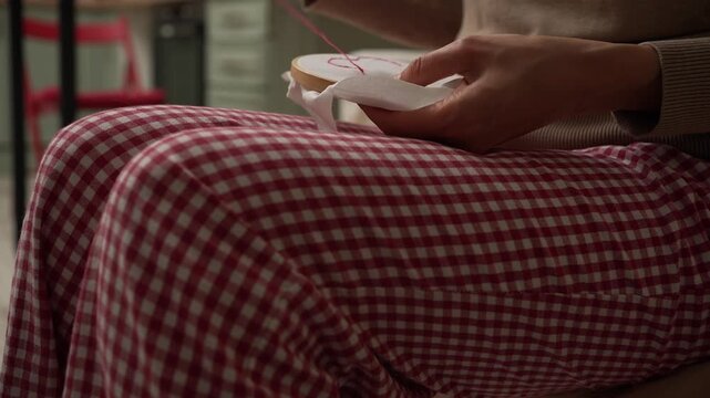 Close-up of a creative woman's hands doing needlework at home, carefully stitching a design on white fabric held in a wooden embroidery hoop for a relaxing, handmade craft project