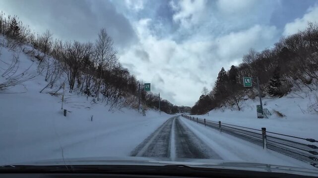 Point of view driving on a winter highway during light snowfall with snow-covered forest and cloudy sky. Calm empty road scene with no traffic