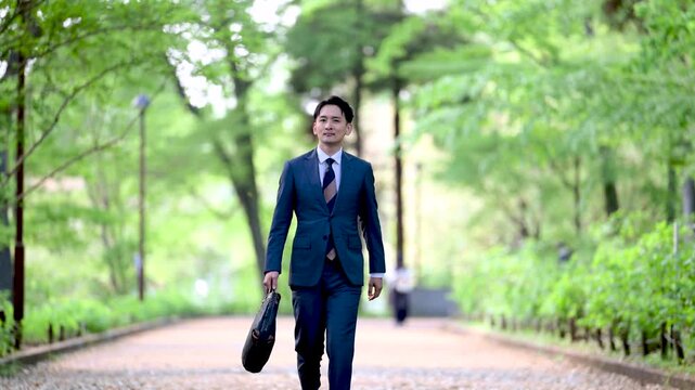 新緑の公園をビジネスバッグを持って歩く哀愁の漂う営業マン仕事イメージの全身の動画スローモーション