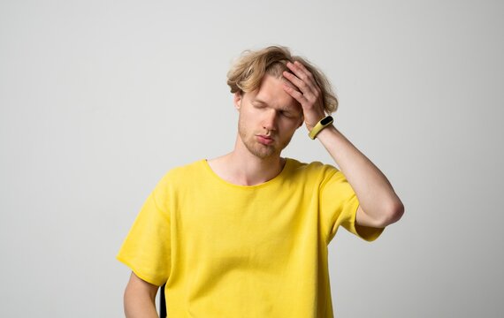 Young adult man experiencing headache with hand on forehead, feeling pain and discomfort, standing against a light background representing stress, fatigue, and illness