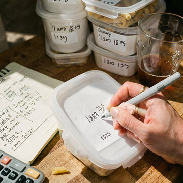 A person writing on a piece of paper next to a plastic container filled with food and a calculator on a wooden table. Freezer meal prep hands.
