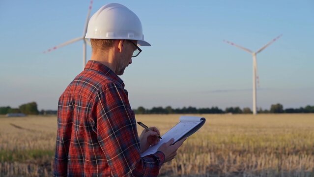 Man engineer, wearing a white protective helmet is taking notes with a clipboard in a field with wind turbines, as the sun sets. Clean energy and engineering concept