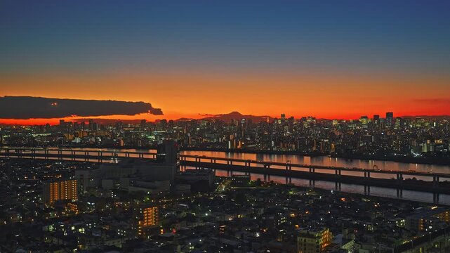 A sweeping drone shot captures vast urban sprawl during a fiery sunset. The camera tracks slowly, highlighting river bridges and a mountain silhouette.