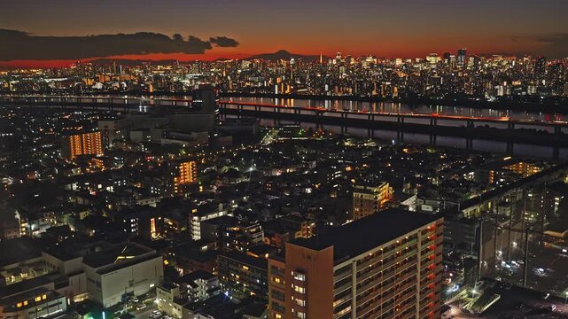 A stunning high-angle shot capturing the Tokyo urban sprawl during the magic hour. The sky glows red as city lights twinkle, with a river and a distant mountain silhouette on the horizon.
