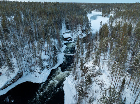 Aerial view of the historic Myllykoski mill and suspension bridge over the Kitkajoki river rapids in a snow-covered forest in Oulanka, Lappi, Finland.