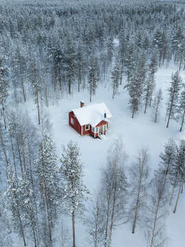 Aerial view of a red wooden cabin surrounded by snow-covered pine trees in a winter forest in Lapland, Finland.