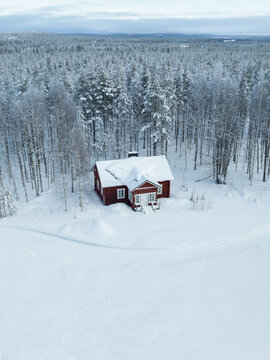 Aerial view of a red wooden cabin surrounded by snow-covered pine trees and a vast winter forest in Lapland, Finland.
