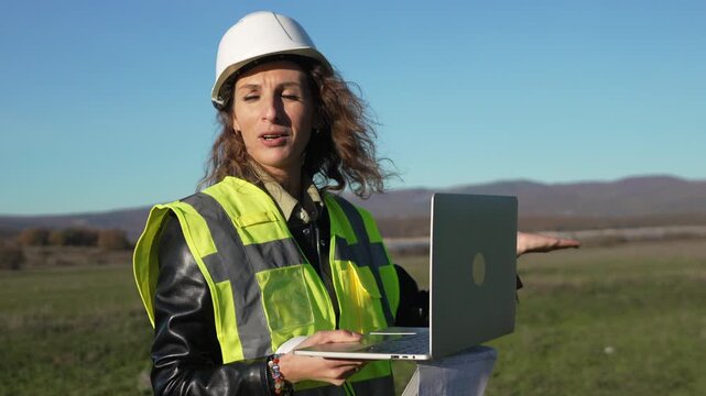 Engineer female construction worker stands outdoors reading blueprints and communicating on a laptop during a site inspection.