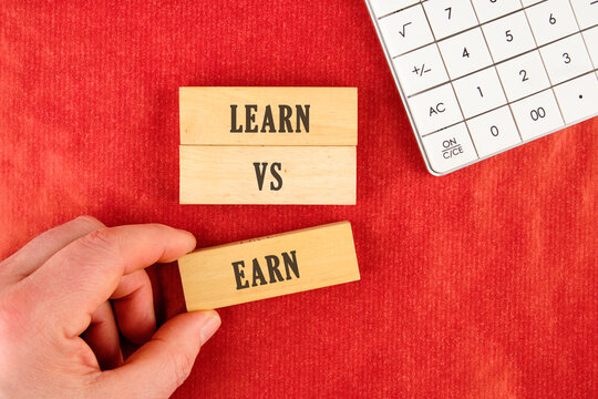 A hand carefully positions a wooden block spelling earn near learn versus blocks. A calculator rests on the bright red textured surface indoors