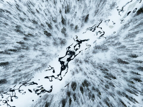 Aerial view of a winding frozen river cutting through a dense snow-covered forest with frost-covered trees in Lapland, Finland.