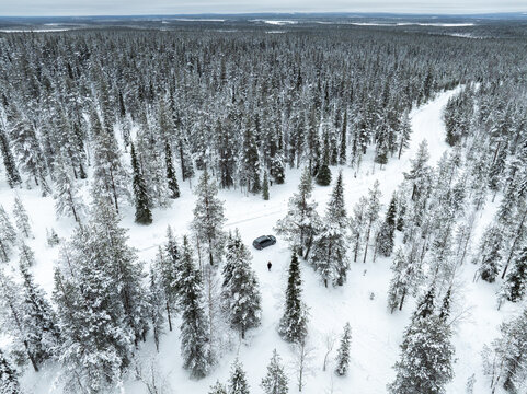 Aerial view of a dark car parked on a snowy path amidst a dense snow-covered pine forest with a person standing nearby in Lapland, Finland.