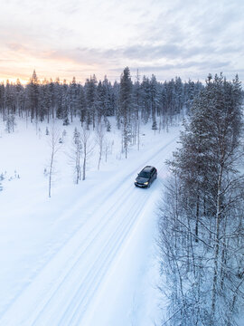 Aerial view of a dark car driving on a snowy road through a dense pine forest during a soft winter sunset Lapland, Finland.