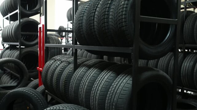 Black rubber tires are organized on metal racks in a tire shop, with more tires visible in the background under bright overhead lighting